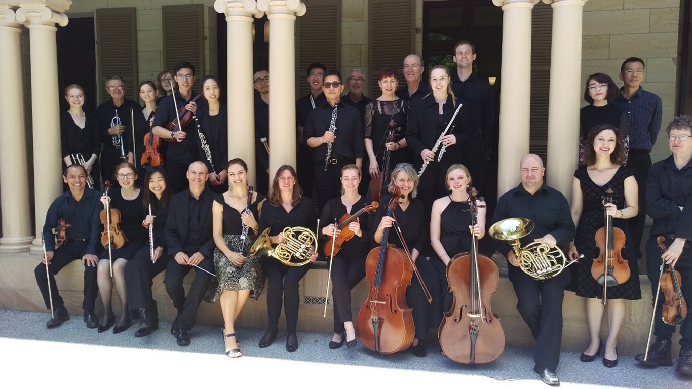 A group of 28 musicians posing for a group photo next to a sandstone building. Half of them are sitting and the other half are standing behind them. They are all holding their instruments